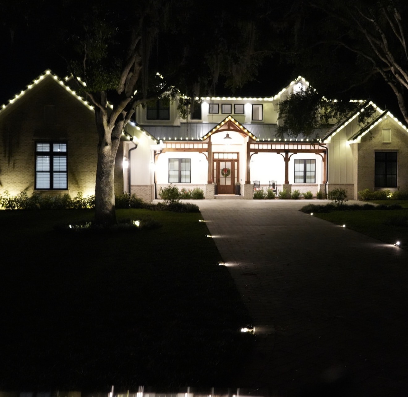 Warm white lights outlining a home beneath mature oak trees